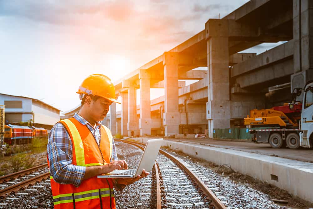 A man holding an iPad, wearing a hard hat and safety vest at a construction site with the sunset in the background