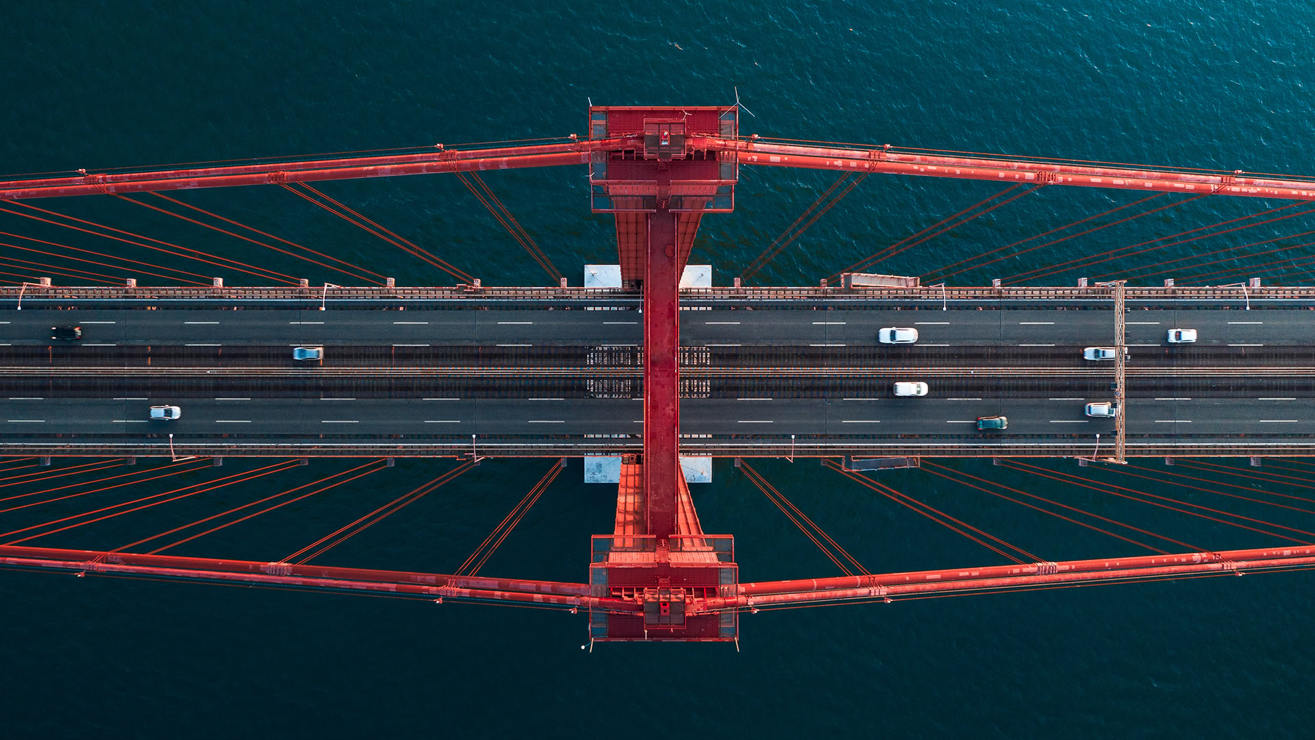 Aerial view of a red suspension bridge in 2024, showcasing the vibrant scene of transportation with multiple vehicles driving on it, set against a backdrop of blue water.
