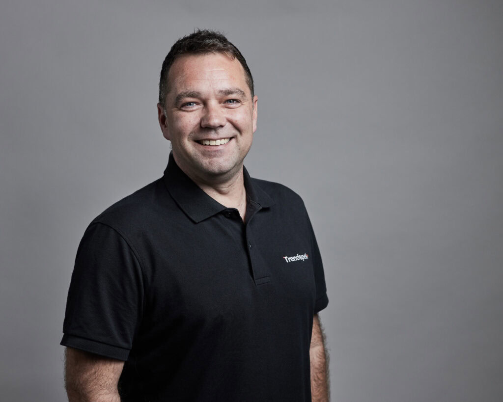 A man in a black polo shirt stands against a plain grey background, smiling at the camera.