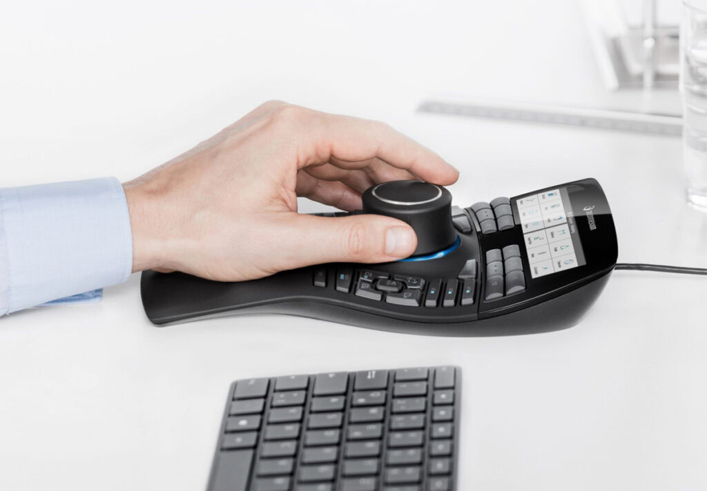 A person uses a 3D navigation controller with an array of buttons and a central control knob while working in MicroStation 2026, next to a computer keyboard on a white desk.
