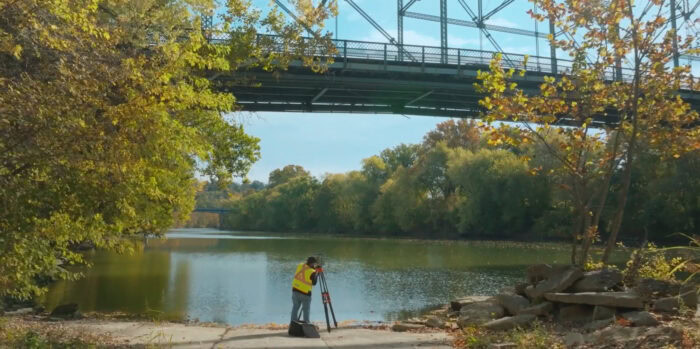 A person wearing a safety vest uses Qk4 surveying equipment near a river, with a steel bridge and trees in the background.