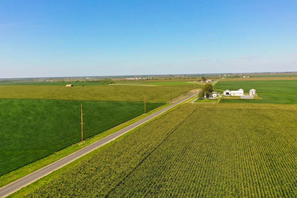 Aerial view of a rural landscape with a paved road dividing green fields, featuring a farmhouse and buildings on the right. Toth and Associates' digital line design enhances infrastructure planning under the clear blue sky.