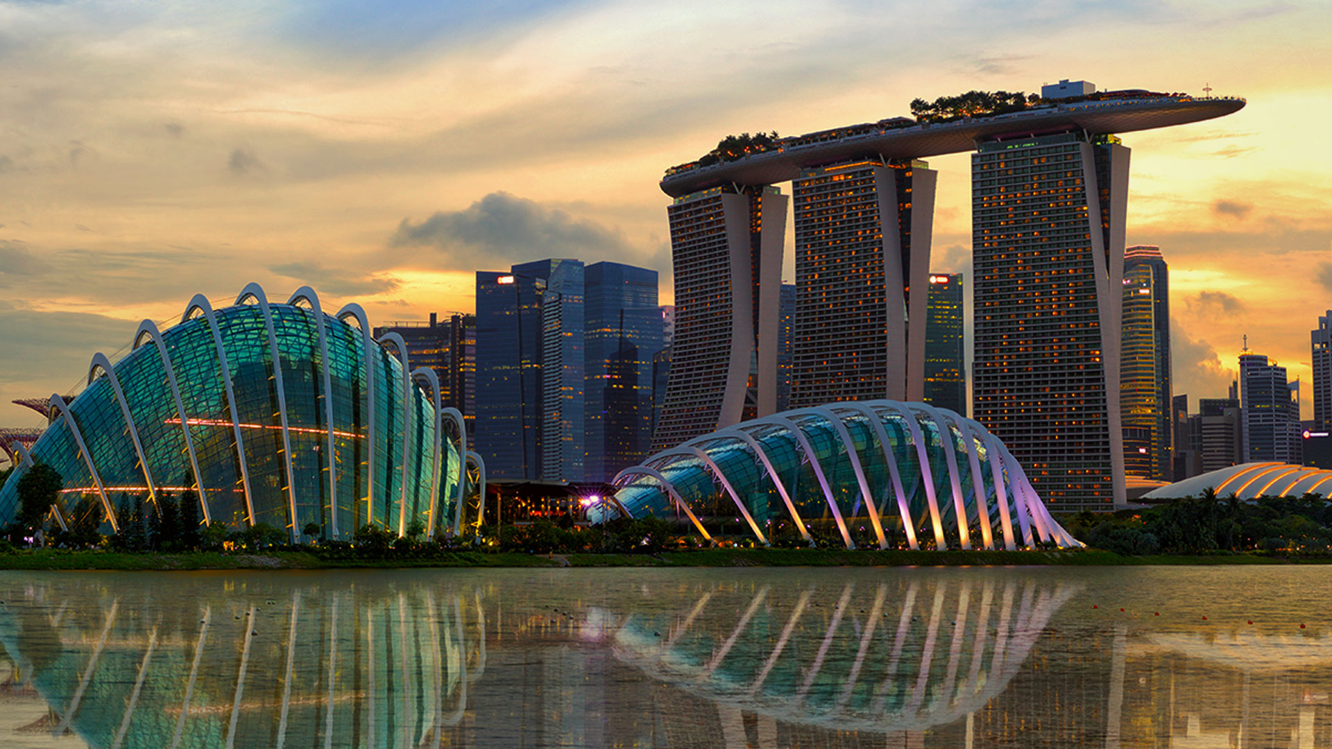 A stunning view of the Marina Bay Sands complex and the Flower Dome in Singapore during sunset, with their reflections visible in the water below. The Marina Bay Sands features three skyscrapers topped by a boat-shaped skypark, while the Flower Dome's illuminated glass structure curves gracefully in the foreground.