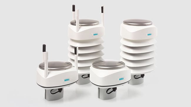 Four white electronic weather sensors with circular tops and varying heights, some with antennas and vented shielding, positioned on a light gray background.