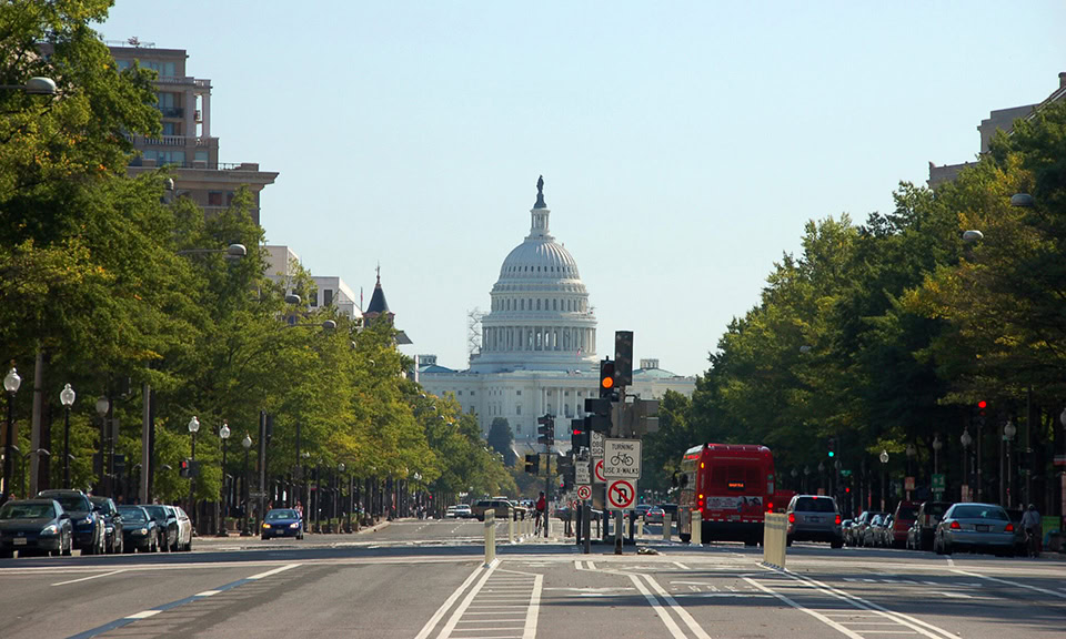 A street view of the United States Capitol building in Washington, D.C., with traffic lights, cars, and trees lining the road.