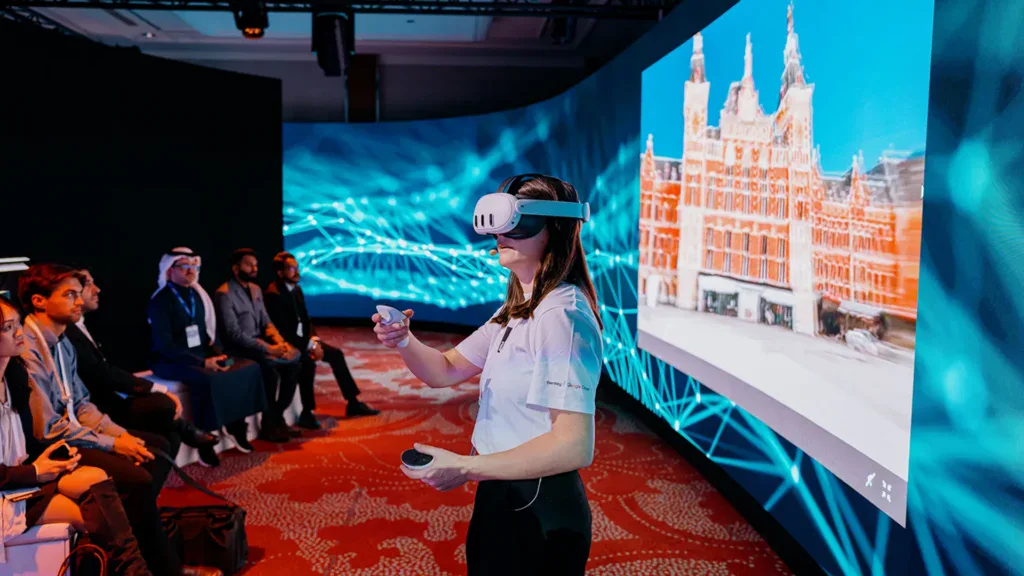 A woman uses a VR headset and controllers in front of a large digital screen showing a building, while an audience watches in a conference setting.