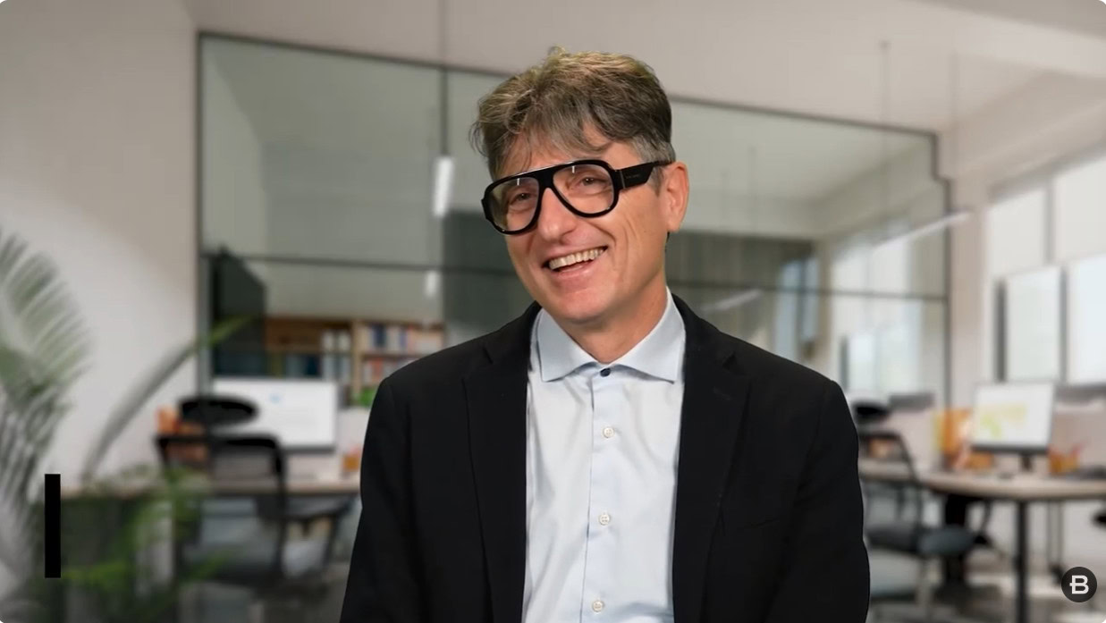 A man wearing black glasses, a light blue shirt, and a dark blazer is smiling while seated in a modern office setting with glass walls and desks in the background for this Planstudio Partner Spotlight.