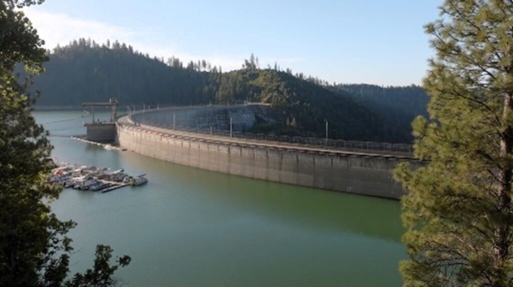 The majestic Bullards Bar Dam spans a verdant lake, nestled amidst forested hills under clear skies, resembling a grand concrete column in nature's vast architecture.